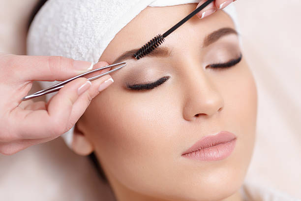 Permanent makeup. Beautiful young woman gets eyebrow correction procedure. Young woman tweezing her eyebrows in beauty saloon. Young woman plucking eyebrows with tweezers close up